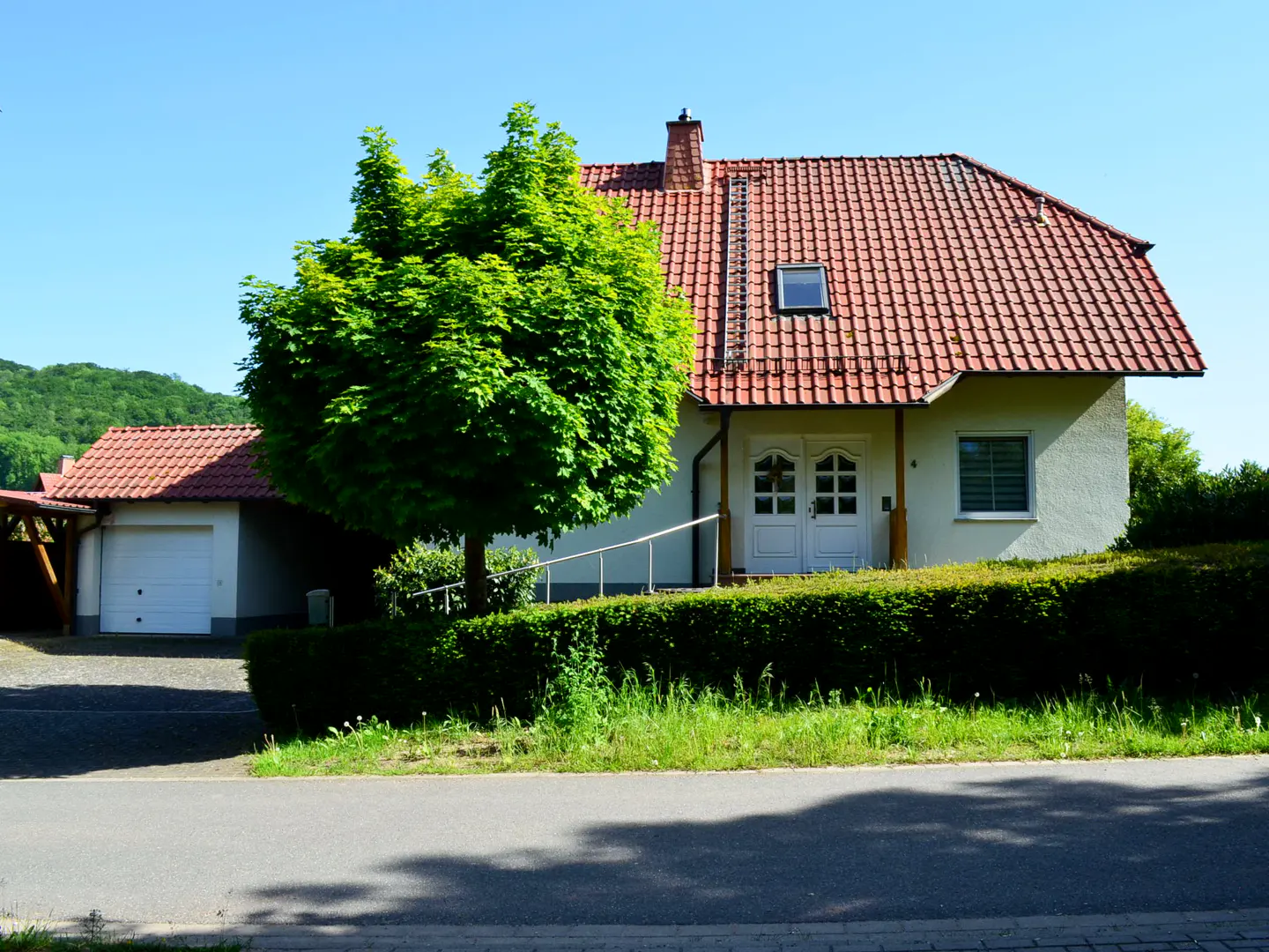 A cream-colored house with a red tile roof, a white garage, and a large green tree in front under a clear blue sky.