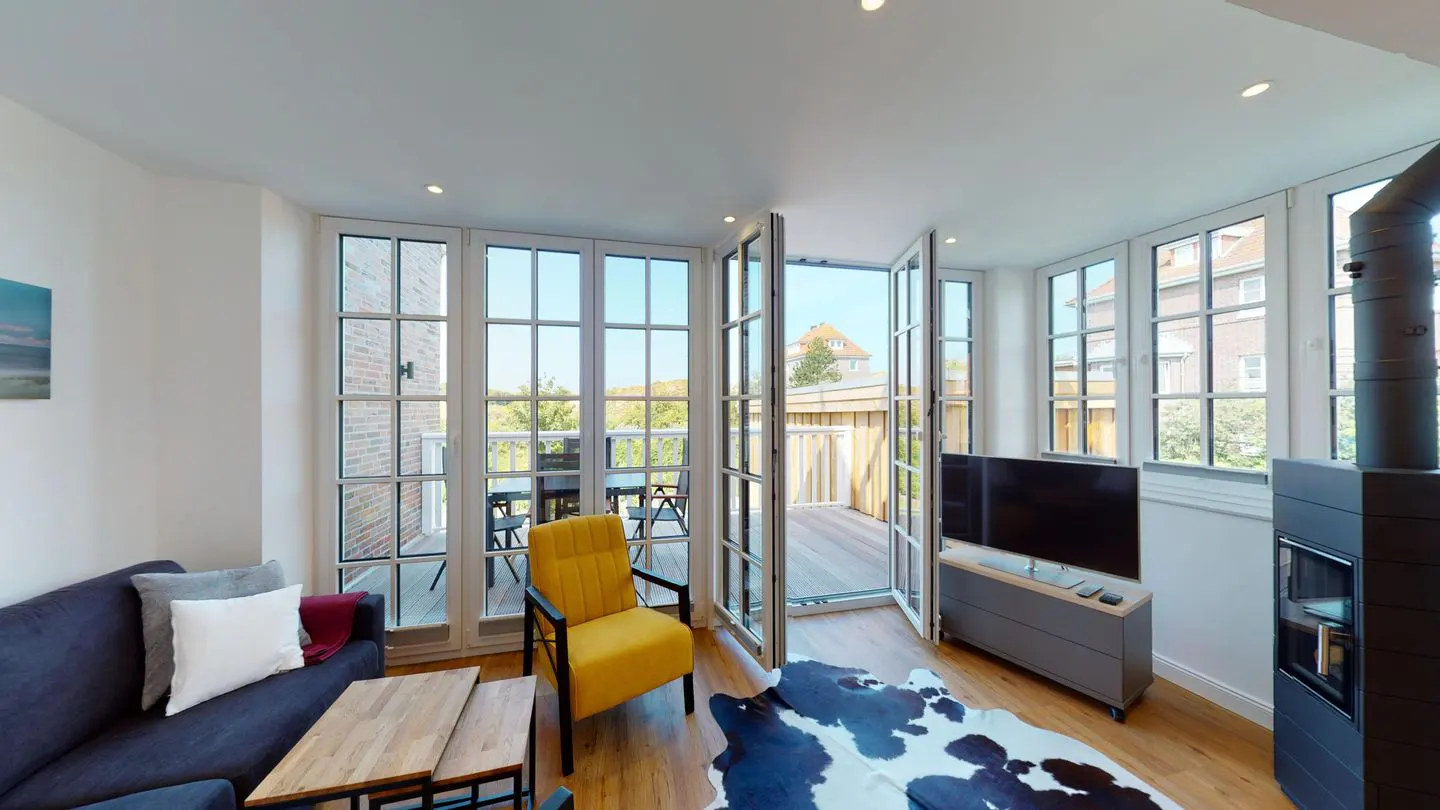Bright living room with blue sofa, yellow chair, and open doors to a balcony. A black and white cowhide rug lies on the wood floor.