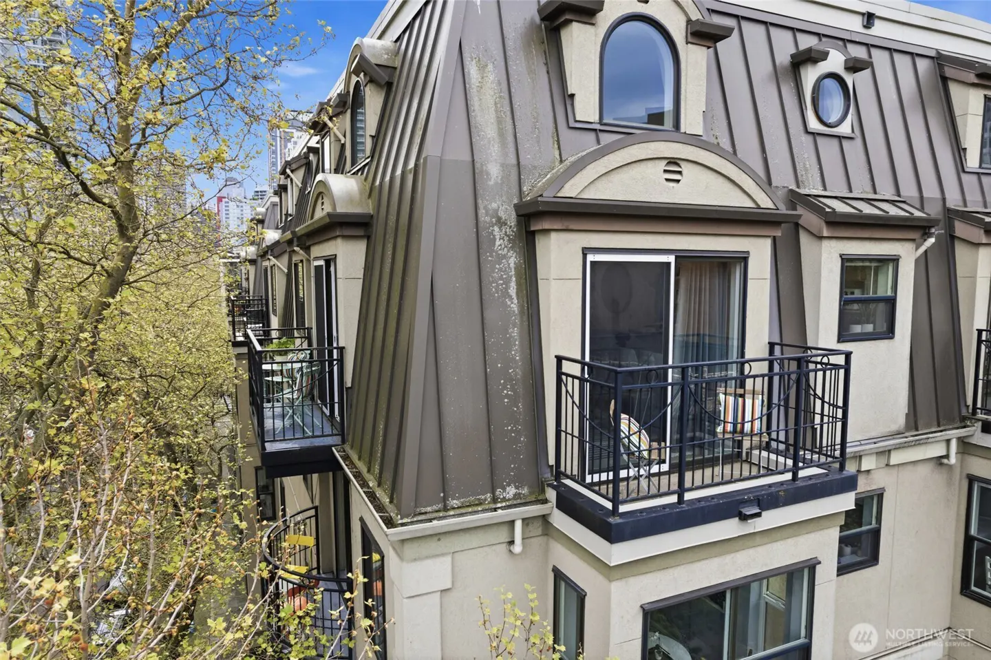 Exterior view of a multi-story building with balconies, gray roof, beige walls, and black railings. A tree with yellow leaves is on the left.