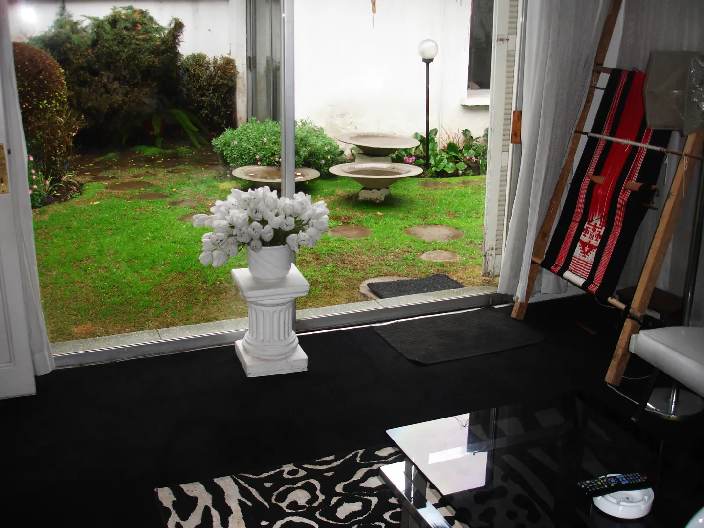Interior view through glass doors to a green garden with bird baths. White tulips sit on a column. Black carpet and zebra rug inside.