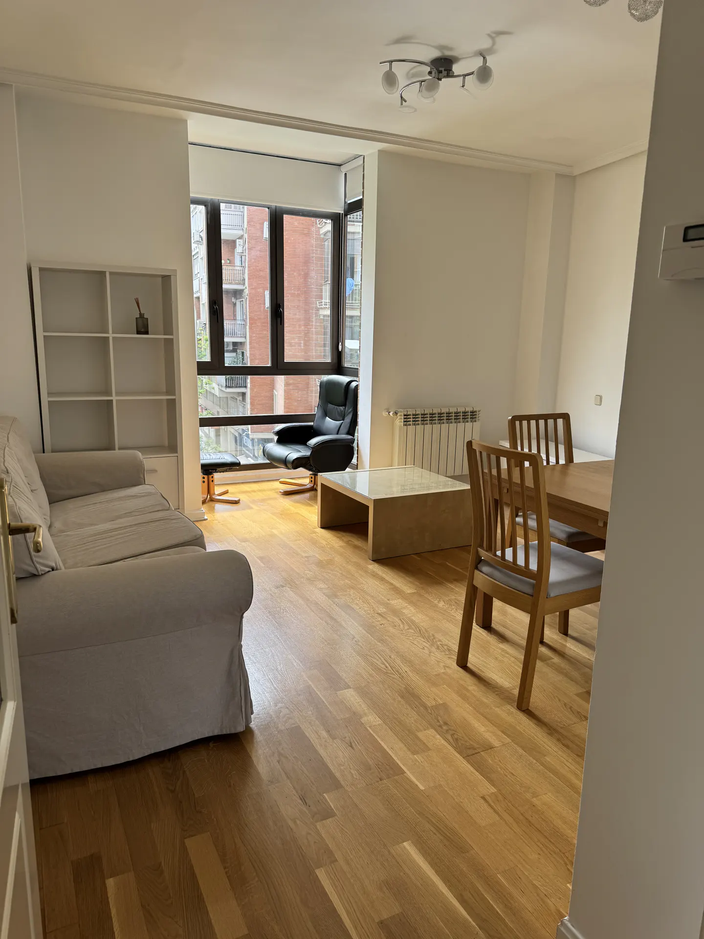 Bright living room with hardwood floors, a beige sofa, a black recliner, and a wooden dining table. Large windows offer a city view.
