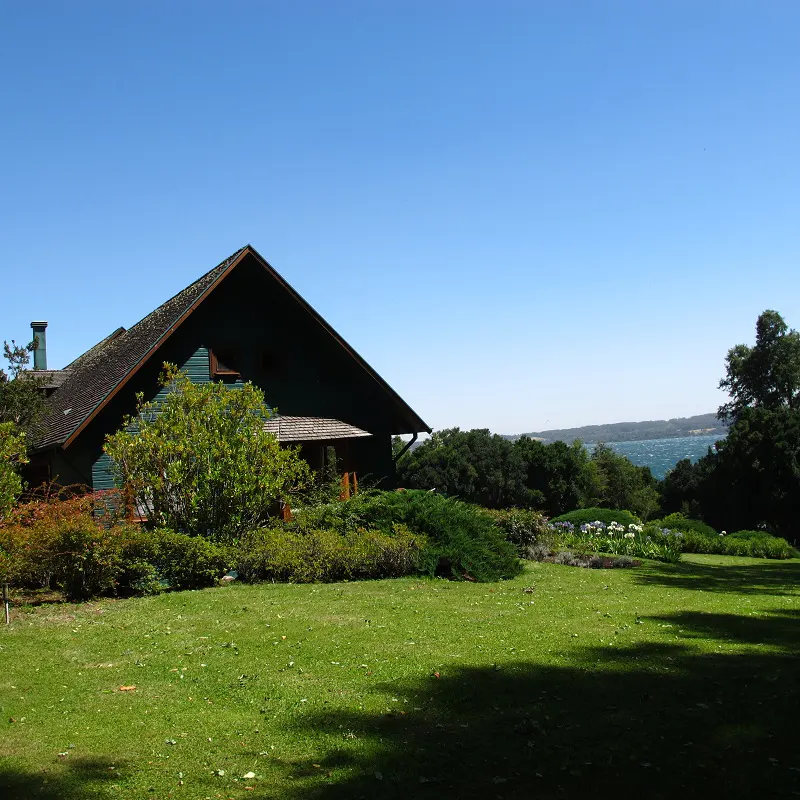 A green house with a brown roof sits on a green lawn under a blue sky, with trees and water in the background.