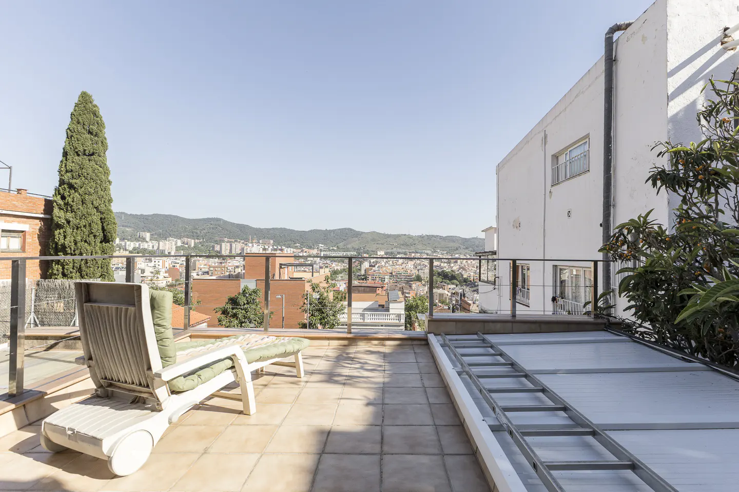 A rooftop terrace with a lounge chair overlooking a city and mountains on a sunny day.