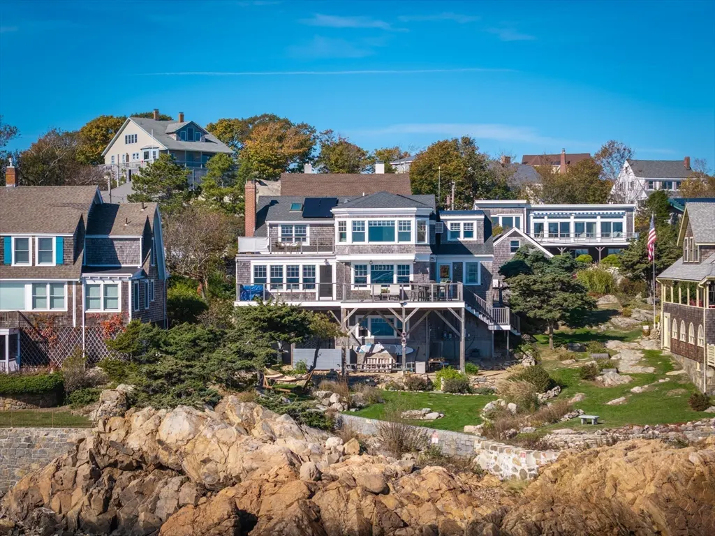 Coastal homes with gray shingles sit on a rocky shoreline under a clear blue sky. Trees and green lawns surround the houses.