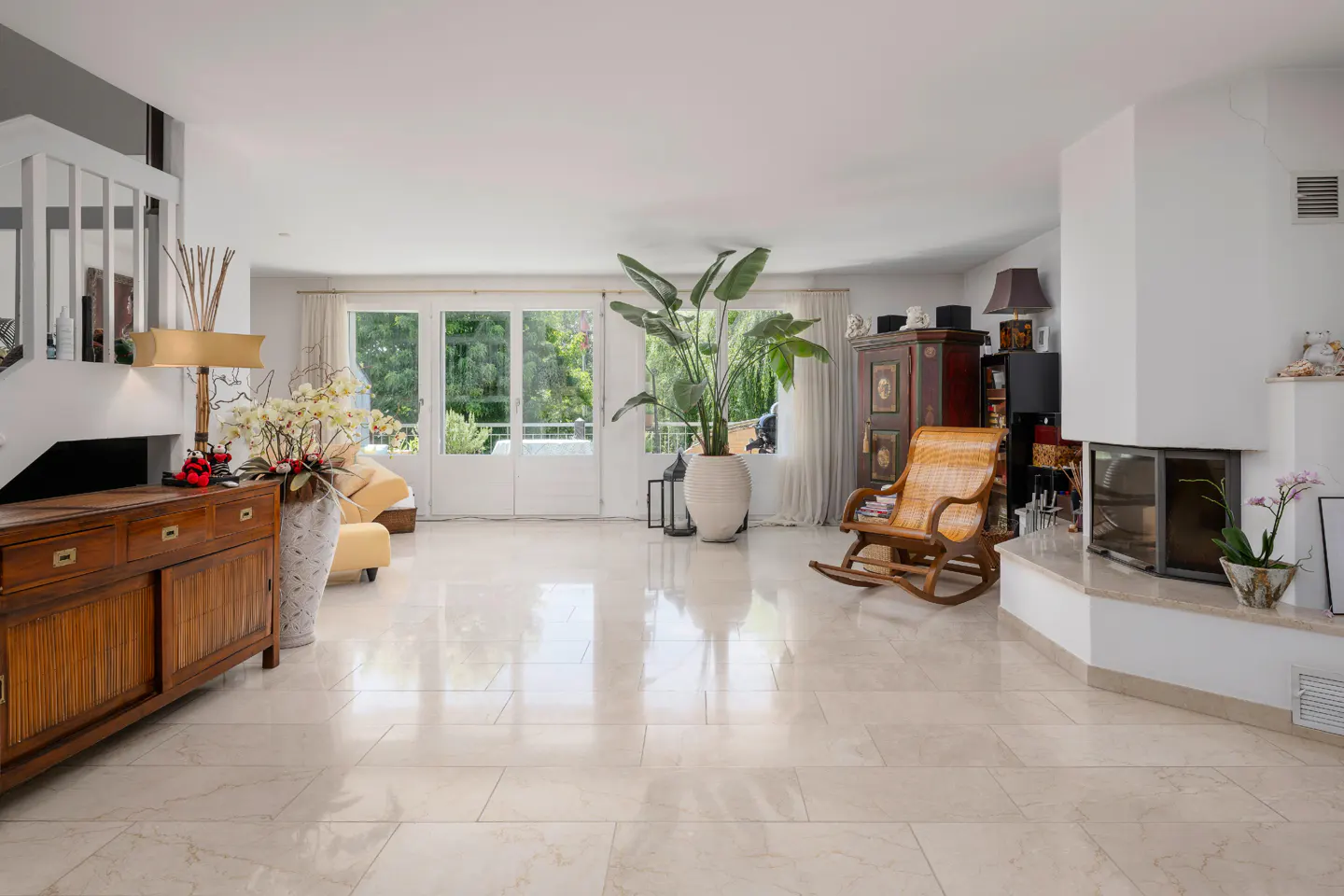 Bright living room with marble floors, white walls, and large windows. A wooden cabinet, rocking chair, and fireplace add warmth to the space.