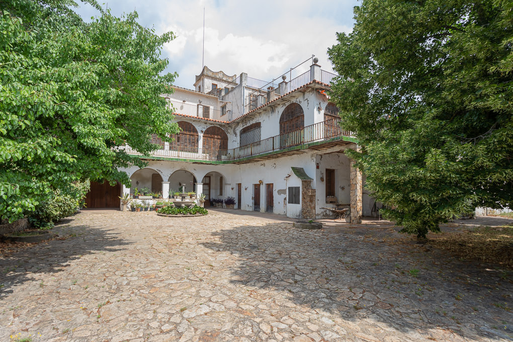 Courtyard view of a white, multi-story building with arched windows and balconies. Stone-paved ground and green trees surround the building.