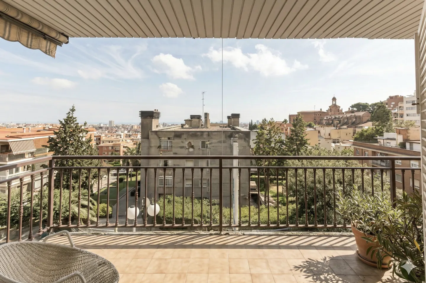 Balcony view with brown railings overlooking a city skyline with trees and buildings under a cloudy sky. A wicker chair and potted plant are on the tiled balcony.