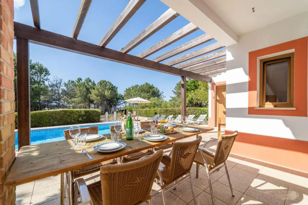 Outdoor dining table set under a wooden pergola on a patio overlooking a swimming pool and lush green trees.