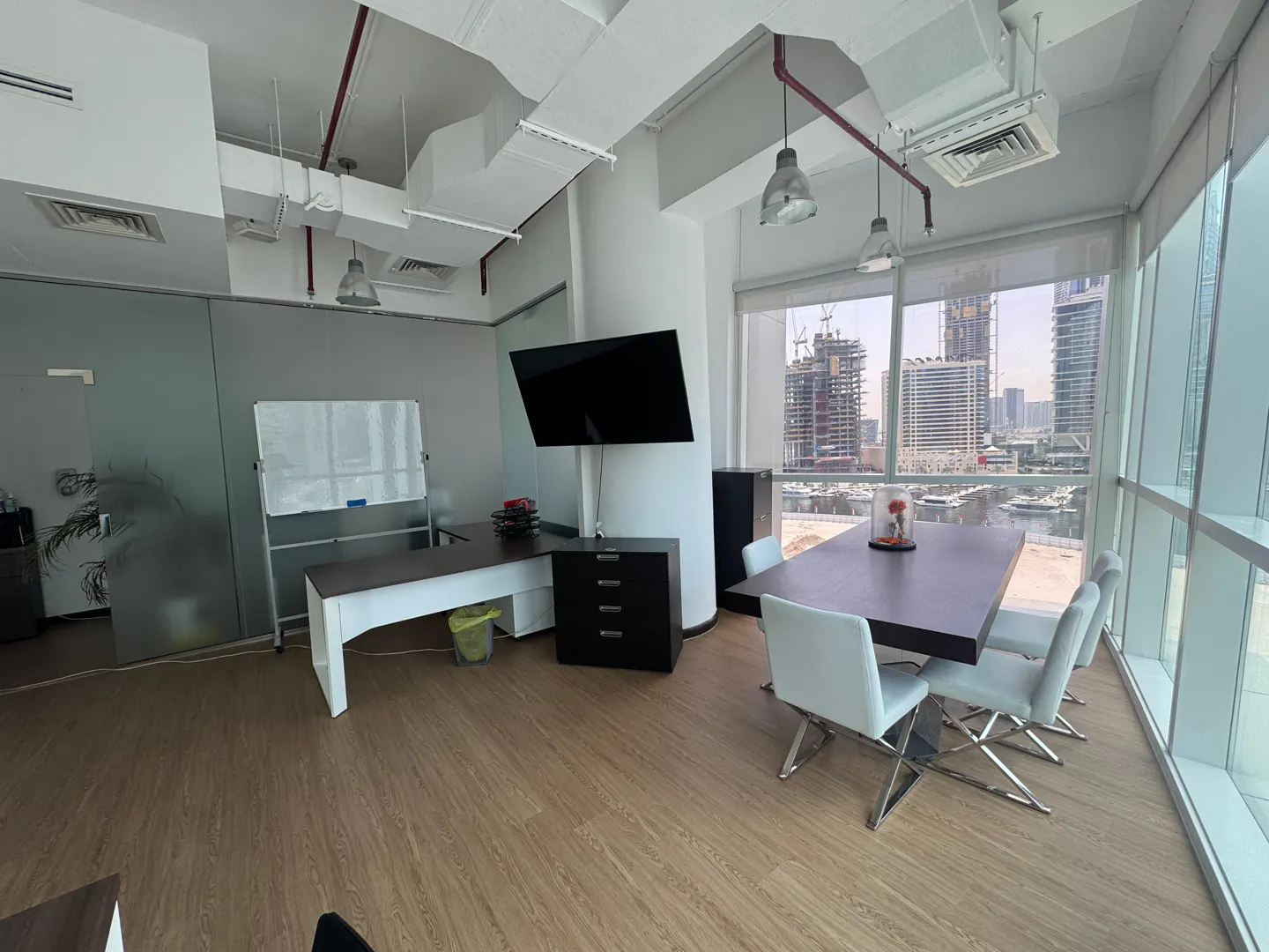 Bright office space with a desk, whiteboard, and a conference table with white chairs. Large windows offer a city view.