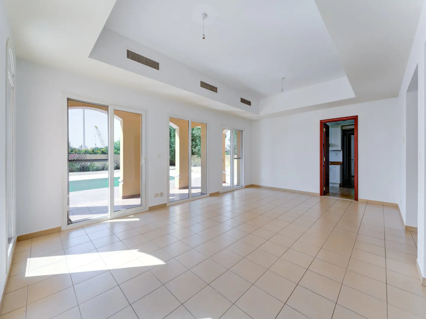 Bright, empty room with white walls, beige tile floor, and sliding glass doors to a pool area. A doorway leads to a kitchen.
