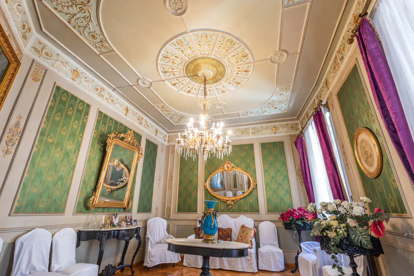 Ornate room with green patterned walls, gold trim, and a decorative ceiling. A chandelier hangs above a table with flowers. Mirrors and purple curtains add elegance.