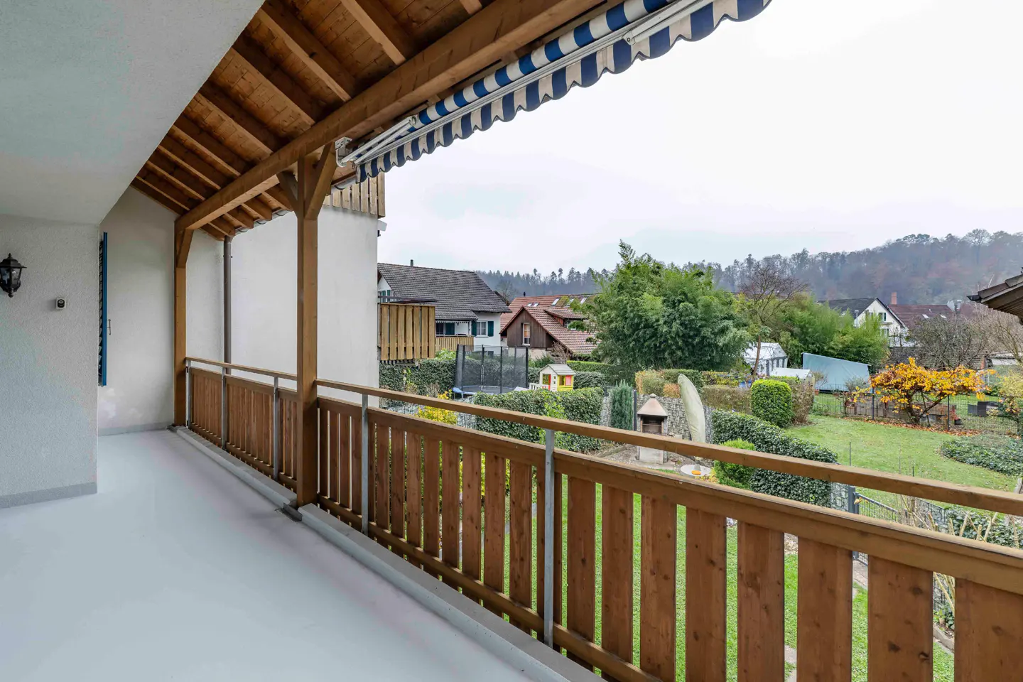 Balcony view with wood railing, awning, and garden. Houses and trees are in the background.