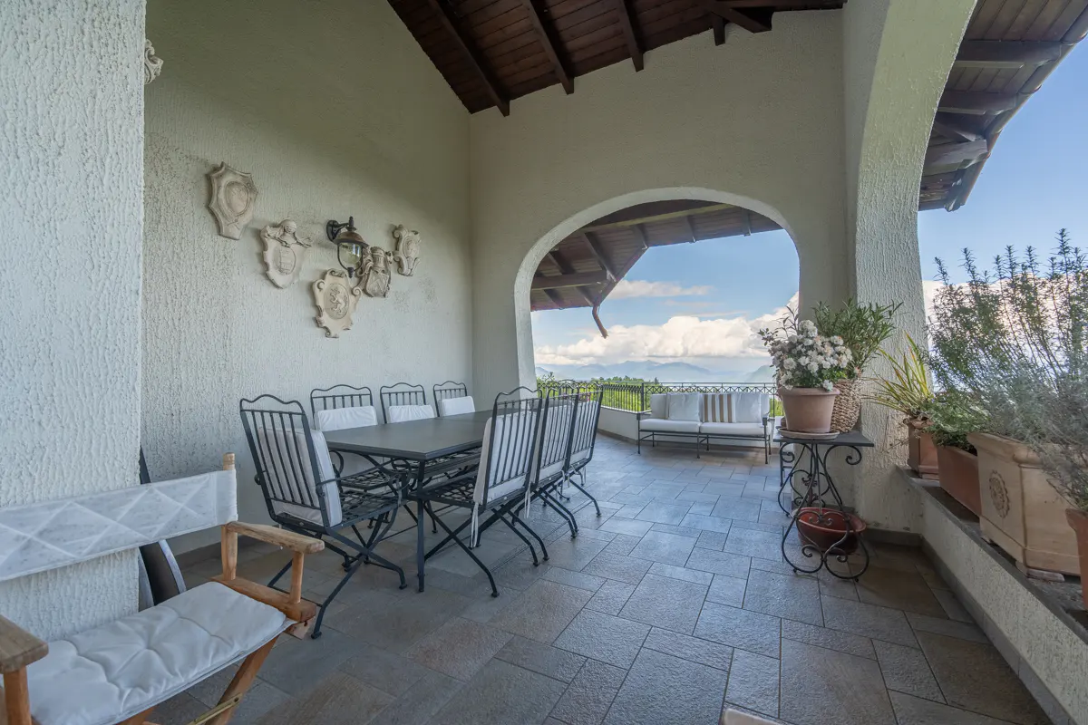 Covered patio with a dining table, chairs, and a view of mountains and sky. Potted plants and a white couch add to the outdoor living space.
