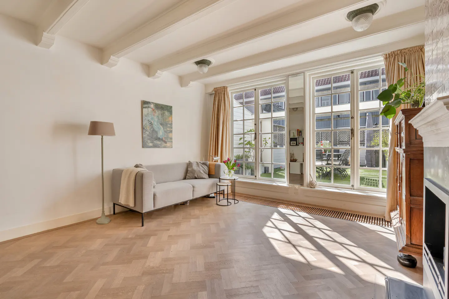 Bright living room with herringbone wood floors, light gray sofa, and large windows overlooking a green backyard.