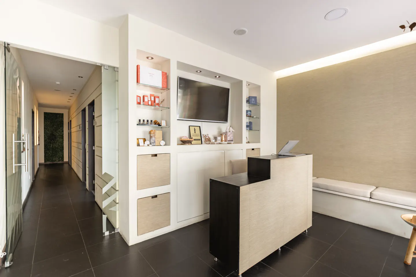 Modern spa reception area with a black and light wood desk, display shelves, and a hallway leading to treatment rooms.