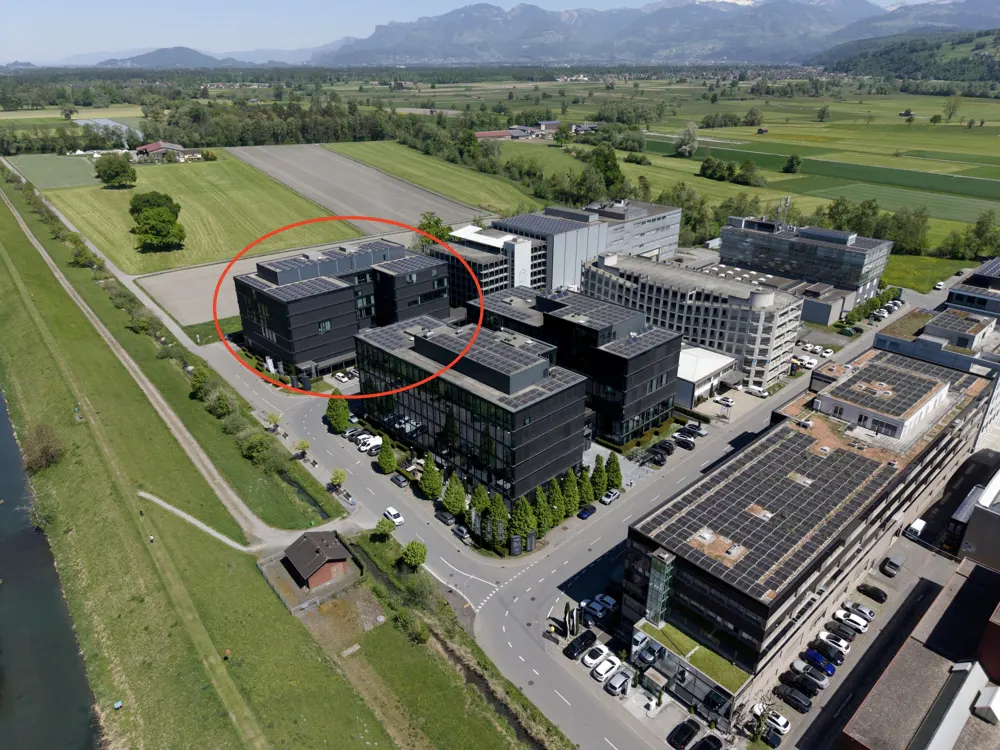 Aerial view of modern black office buildings with solar panels, surrounded by green fields and mountains in the background.