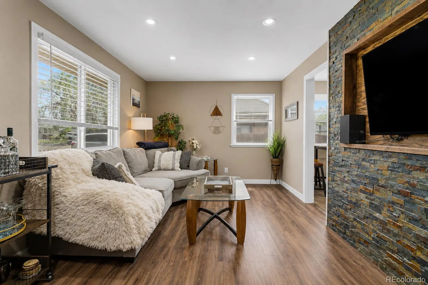 Living room with a gray sectional sofa, glass coffee table, wood floors, and a stone accent wall with a TV.