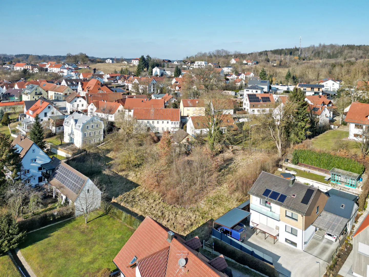 Aerial view of a European village with red-roofed houses, bare trees, and a vacant lot in the foreground. Solar panels are visible on some roofs.