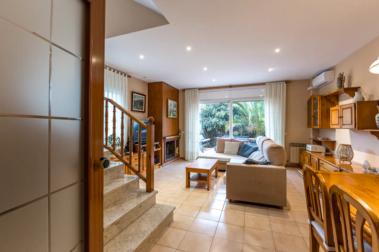 A bright living room with a beige sofa, wooden furniture, and stairs leading up. A glass door opens to the space.