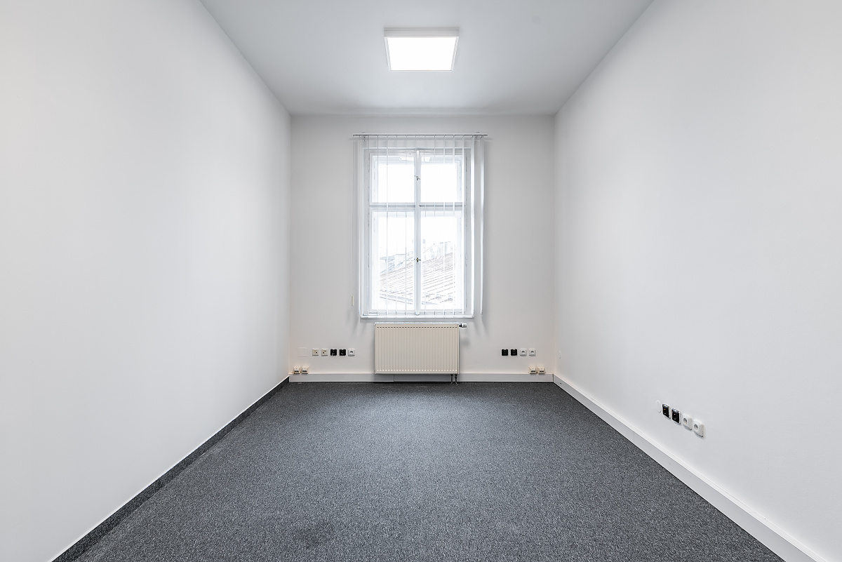 Empty room with gray carpet, white walls, and a window with white curtains. A radiator is under the window. [AI-generated text]