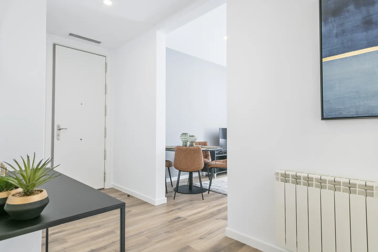 Bright apartment entryway with a white door, black console table with plants, and a glimpse of a dining area with brown chairs.