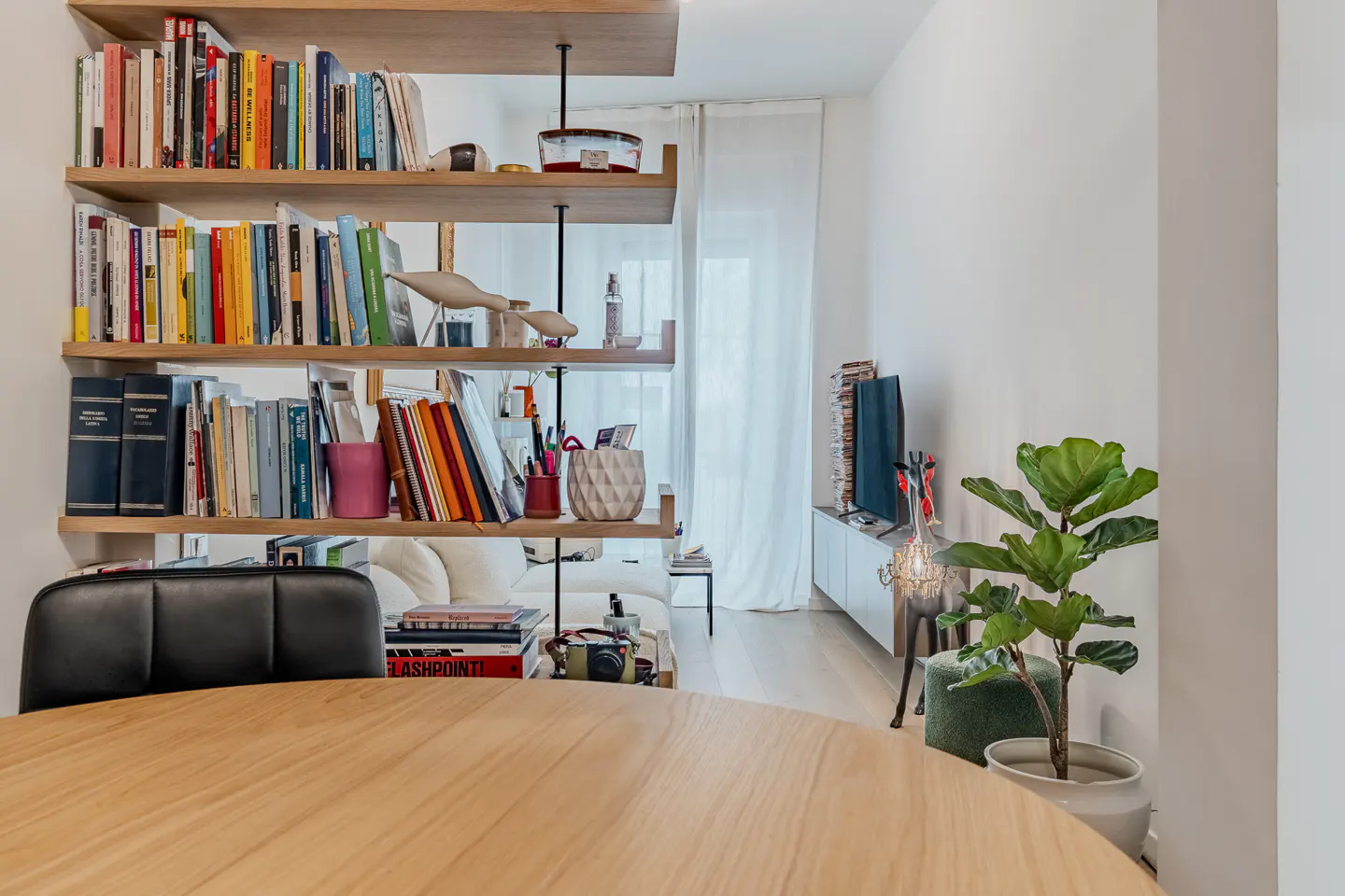 A wooden dining table in the foreground with a floor-to-ceiling bookshelf room divider and a potted plant in the background.