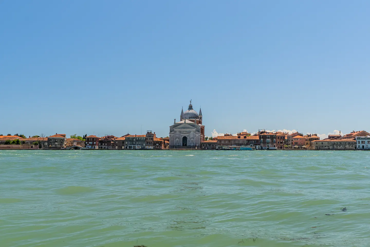 Venice cityscape view with a church and buildings with red roofs, separated by green water under a clear blue sky.