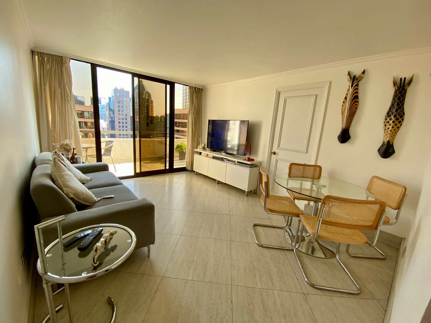 Bright living room with a gray sofa, glass table, and a dining set with wicker chairs. A TV sits on a white cabinet, and zebra masks hang on the wall. Balcony with city views.