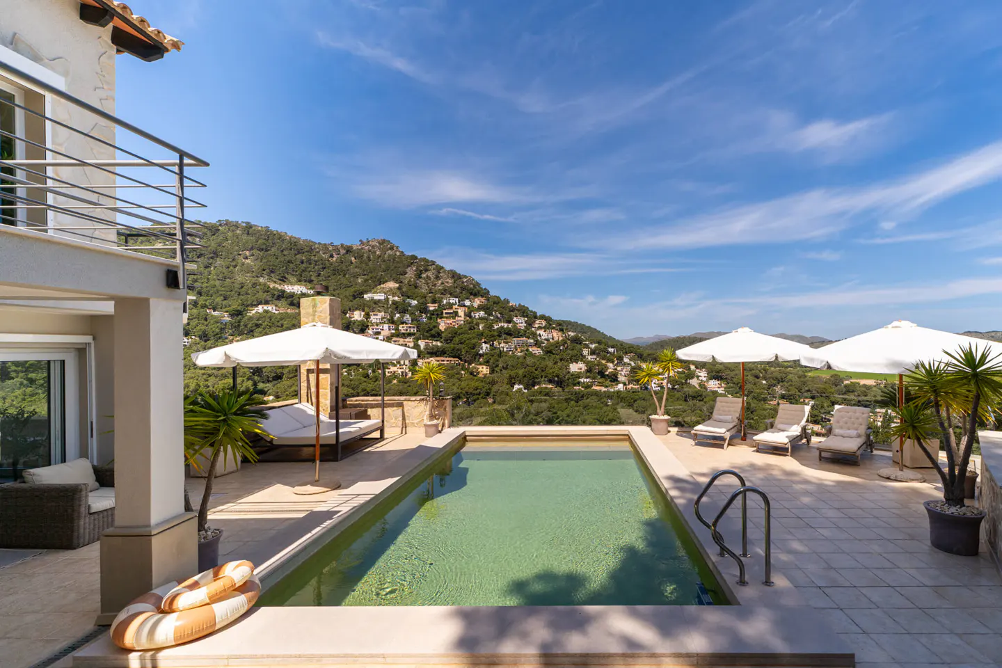 Outdoor pool with lounge chairs and umbrellas on a sunny day. A mountain with houses is in the background.