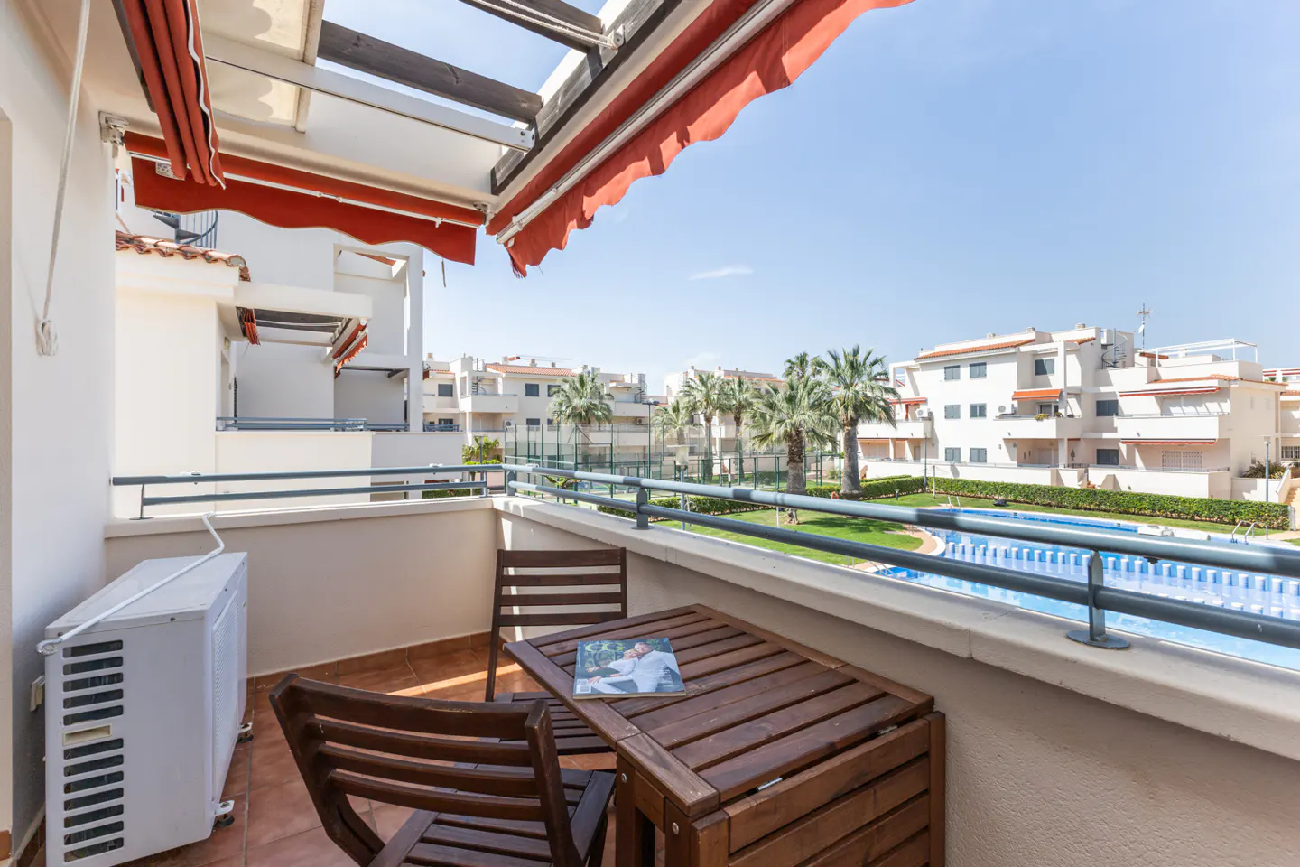 Balcony view with a wooden table and chairs, overlooking a pool and palm trees on a sunny day. Red awning above.
