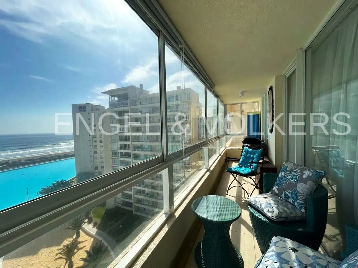 Balcony view with blue chairs, table, and pillows overlooking a large pool, ocean, and buildings under a partly cloudy sky.