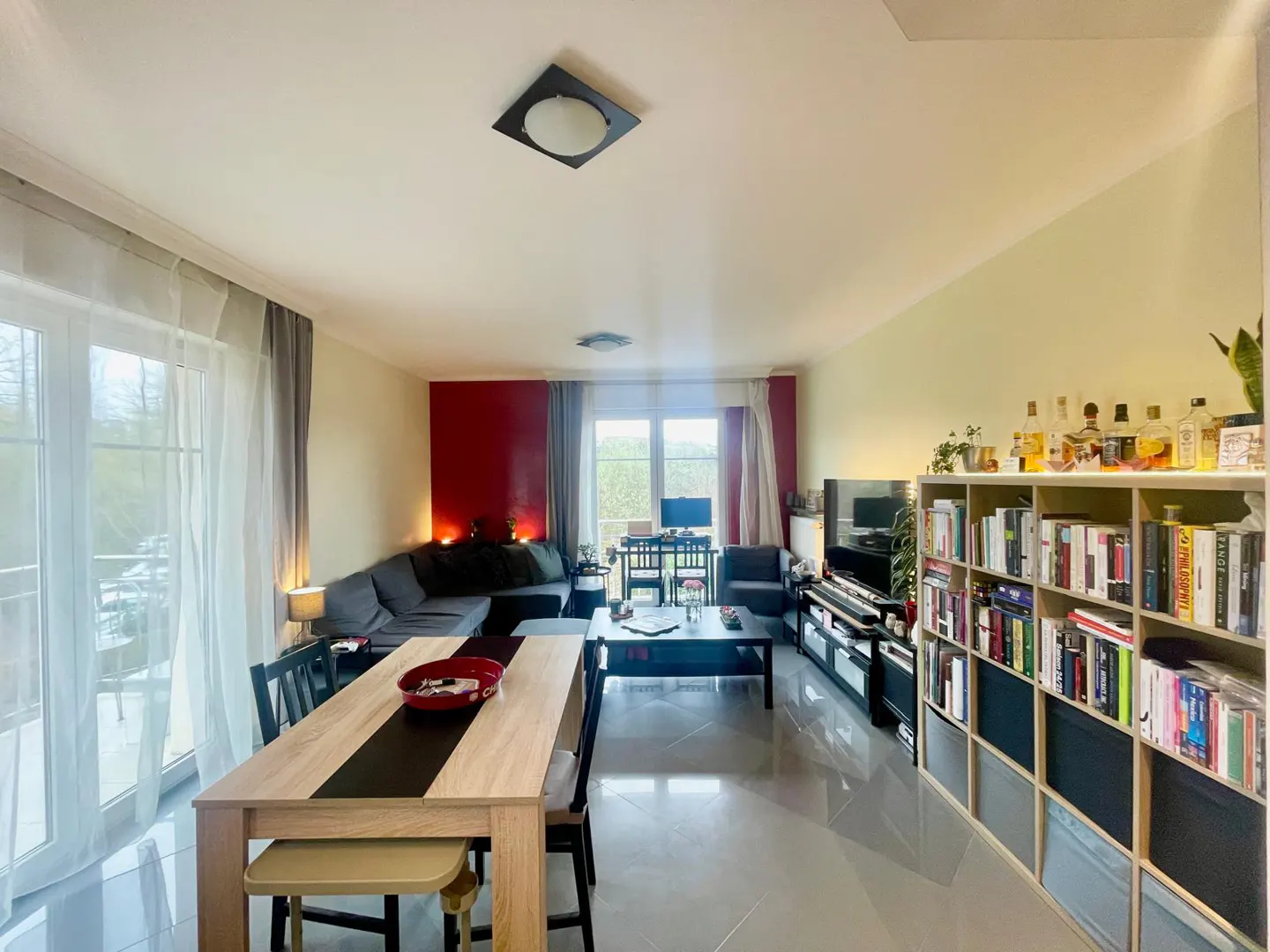 Bright, open living space with a dining table, gray sofa, and a bookshelf filled with books and liquor bottles. Red accent wall.