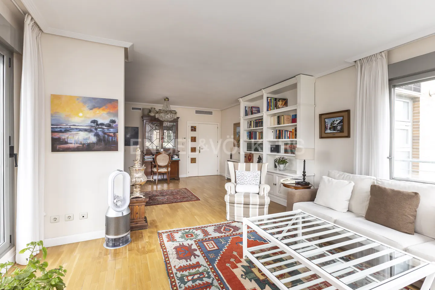 Bright living room with hardwood floors, white sofa, striped armchair, and a glass-topped coffee table over a colorful rug. Bookshelves line the wall.