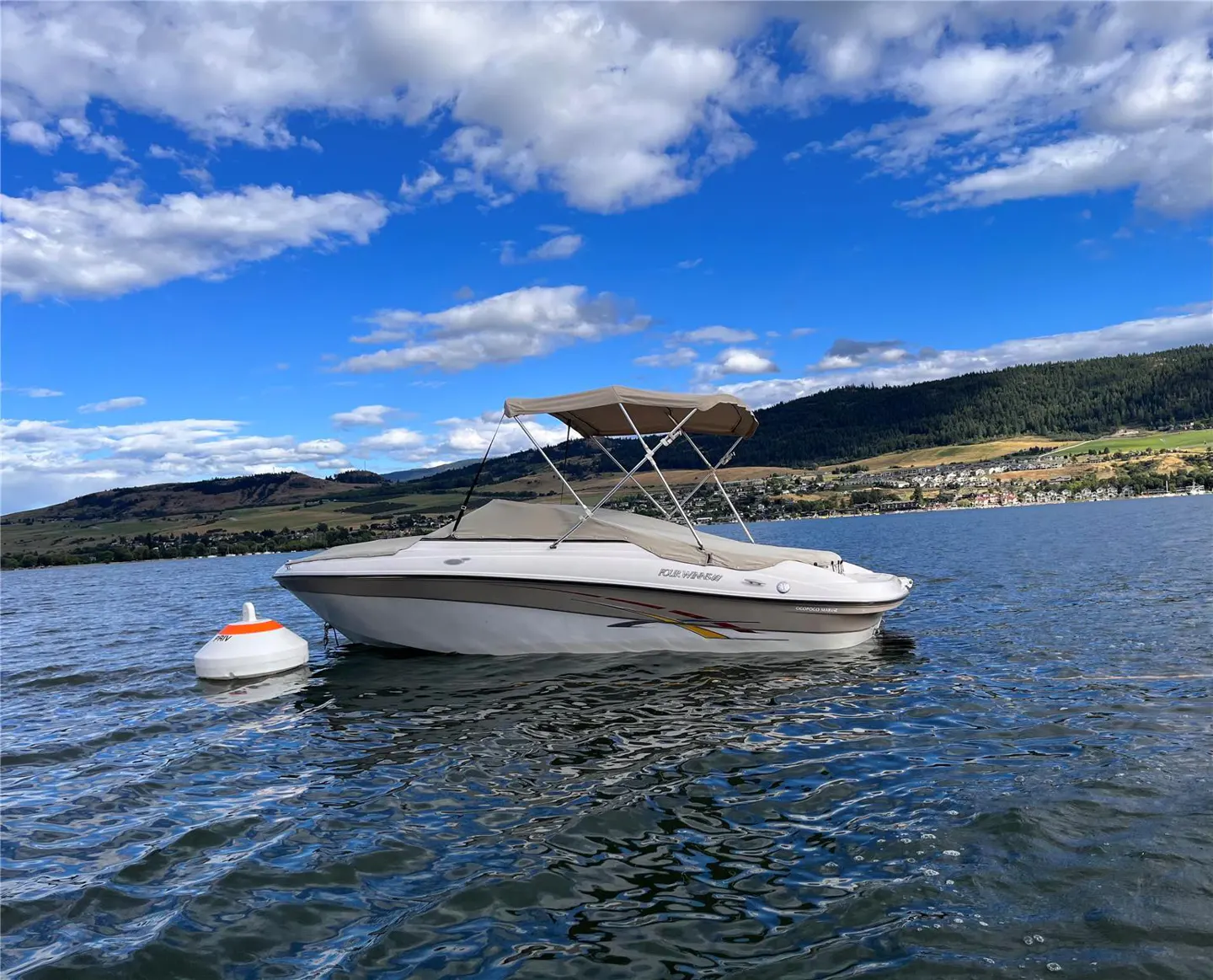 A white and tan boat with a beige canopy sits on a lake under a blue sky with white clouds.
