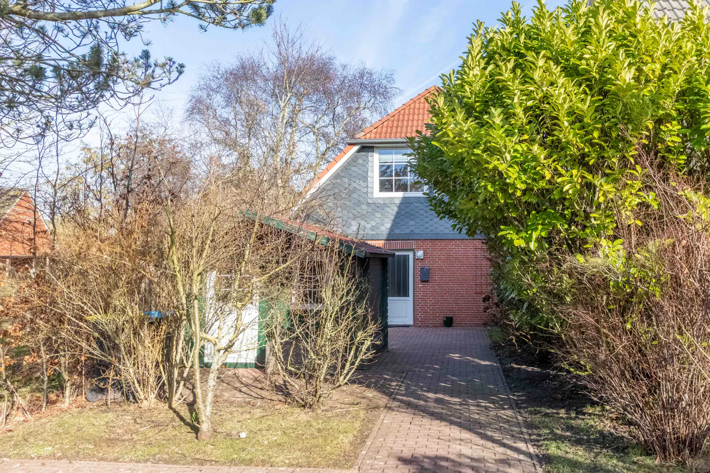 Exterior view of a brick house with a gray roof, white door, and brick walkway, surrounded by trees and bushes.