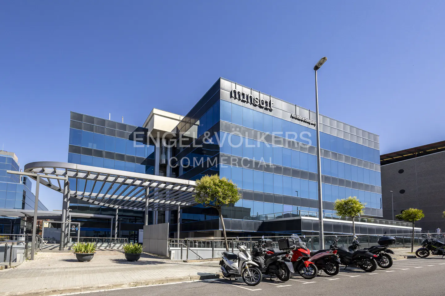 Modern glass office building with a metal pergola, parked scooters, and small trees under a clear blue sky.