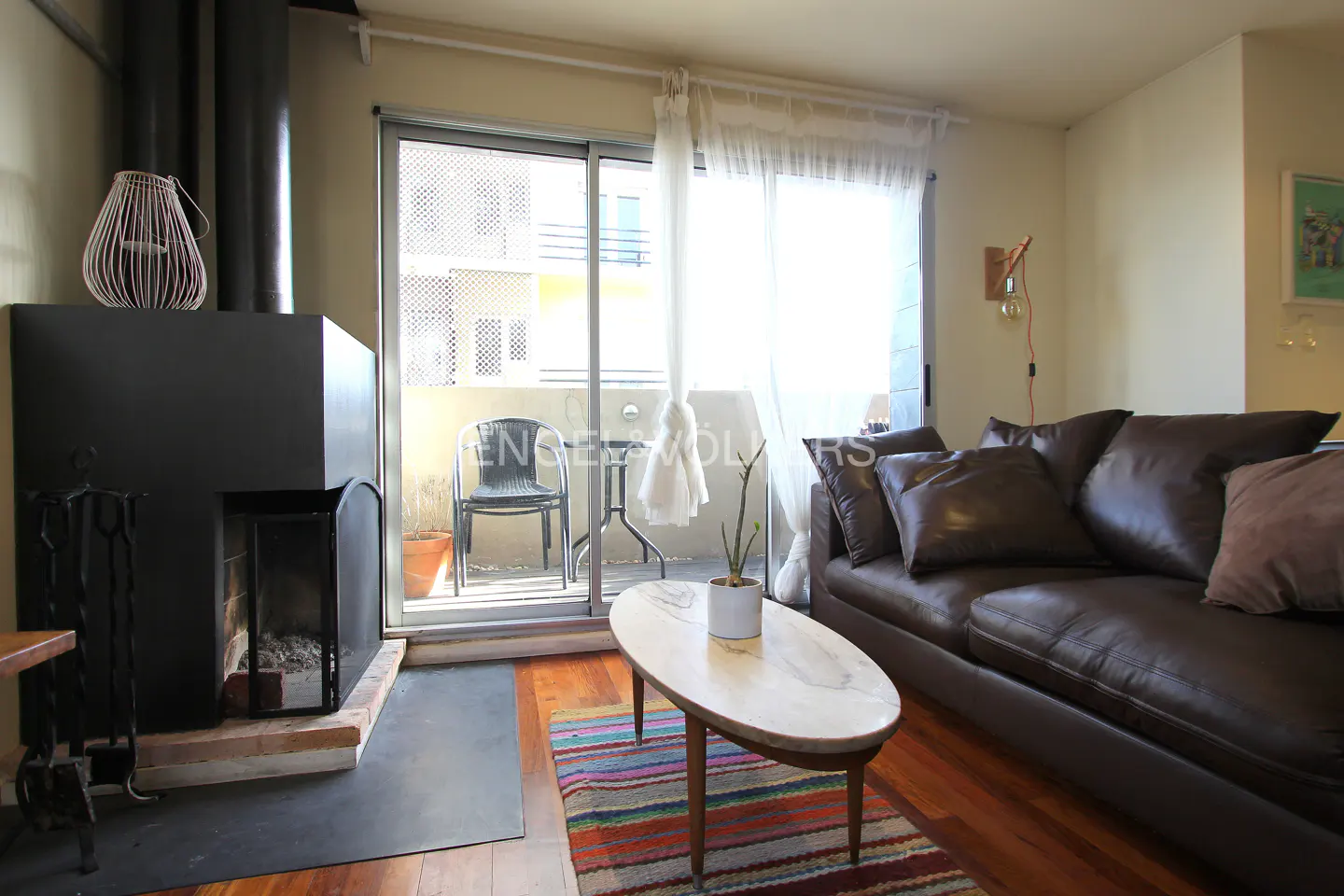 Living room with a black fireplace, brown leather sofa, and a balcony with a chair and table. A colorful rug is on the wood floor.