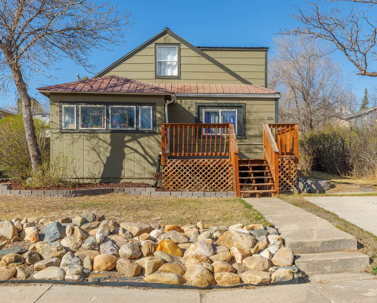 A quaint, light-green house with a brown roof, wooden porch, and rock landscaping under a clear blue sky.