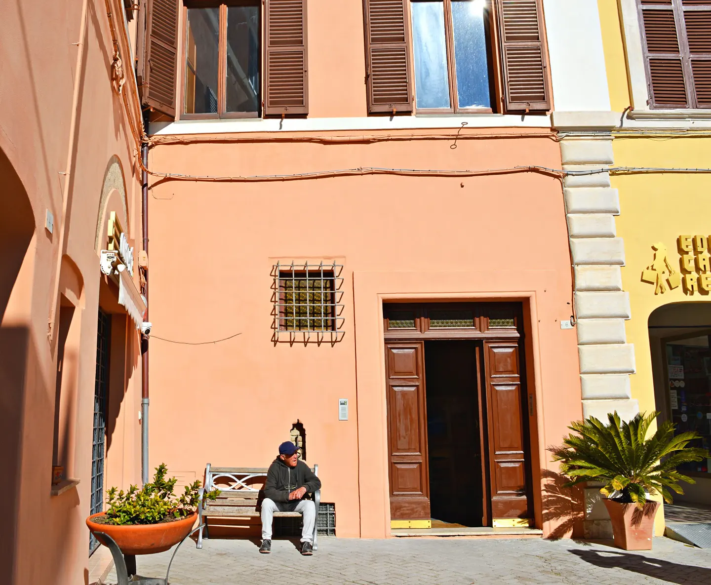 A man sits on a bench in front of a peach-colored building with brown shutters and an open brown door. A potted plant sits nearby.