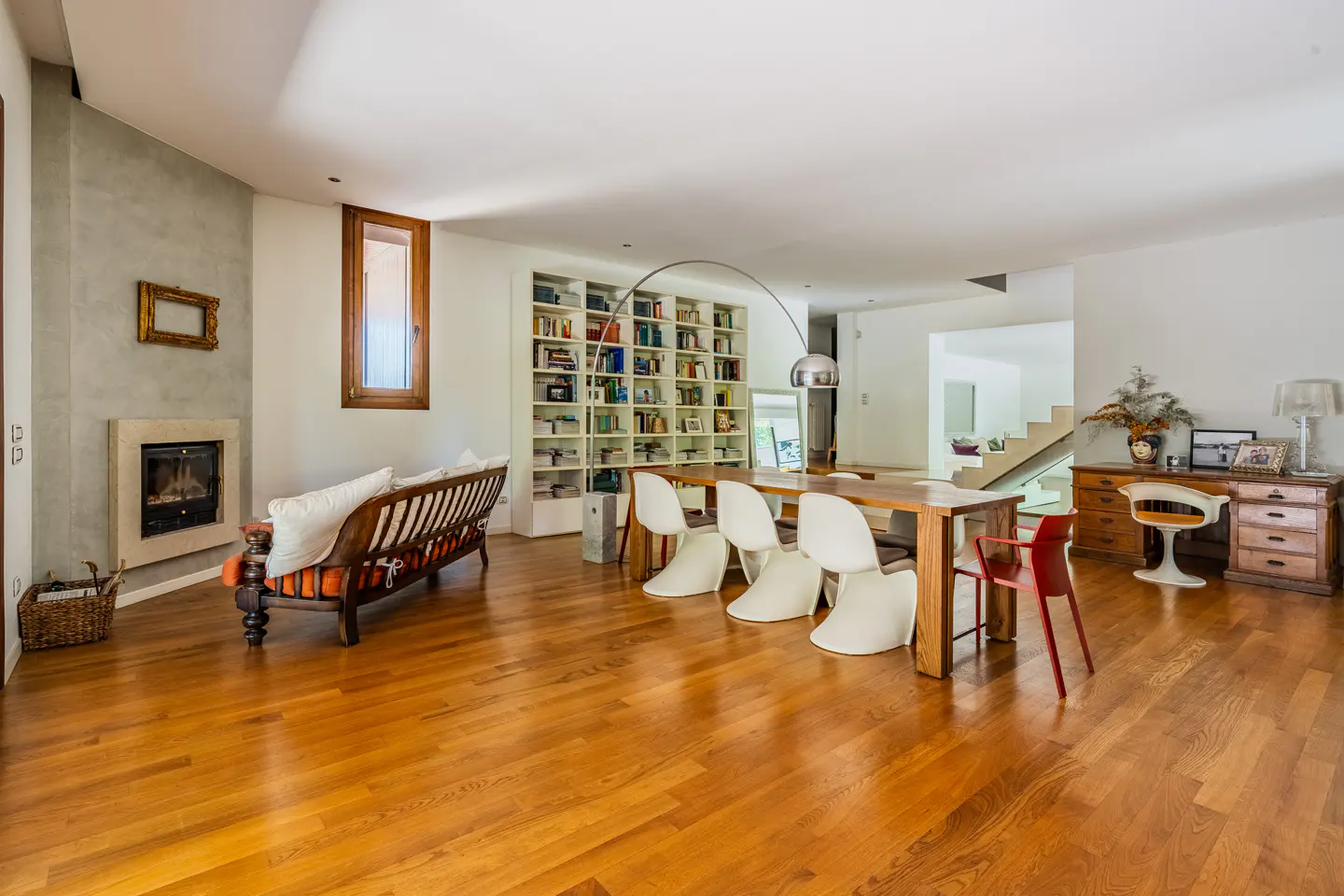Bright, open-plan living room with wood floors, fireplace, sofa, dining table with white chairs, and a large bookcase.