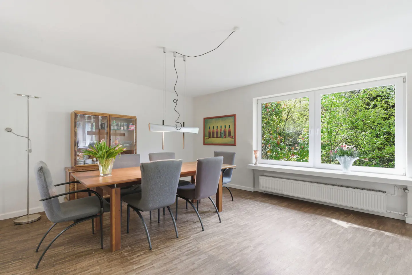 Bright dining room with wood table, gray chairs, and large window overlooking green trees. A glass cabinet and floor lamp are visible.