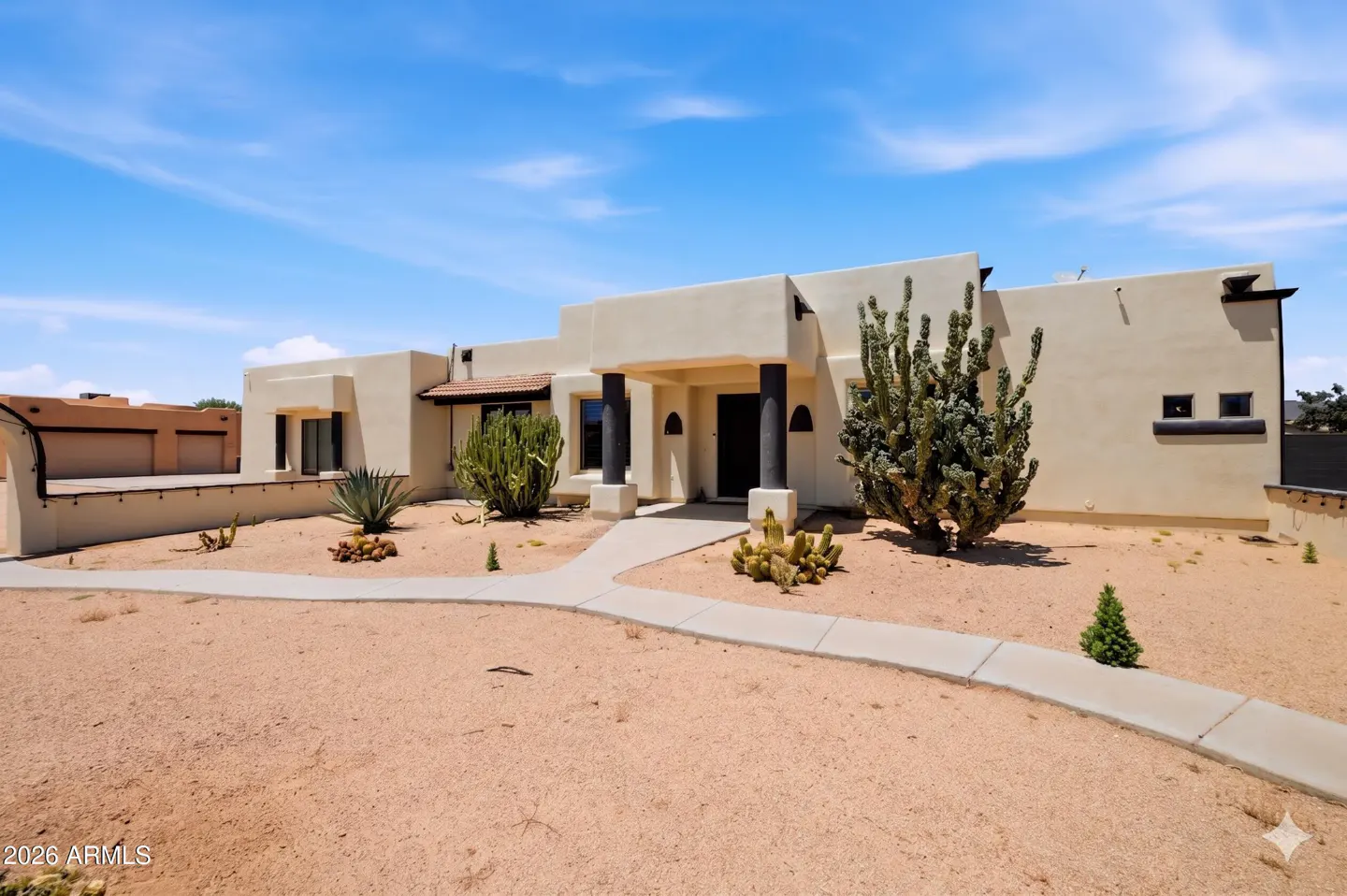 Single-story beige stucco home with a desert landscape, cactus garden, and a concrete walkway under a bright blue sky.