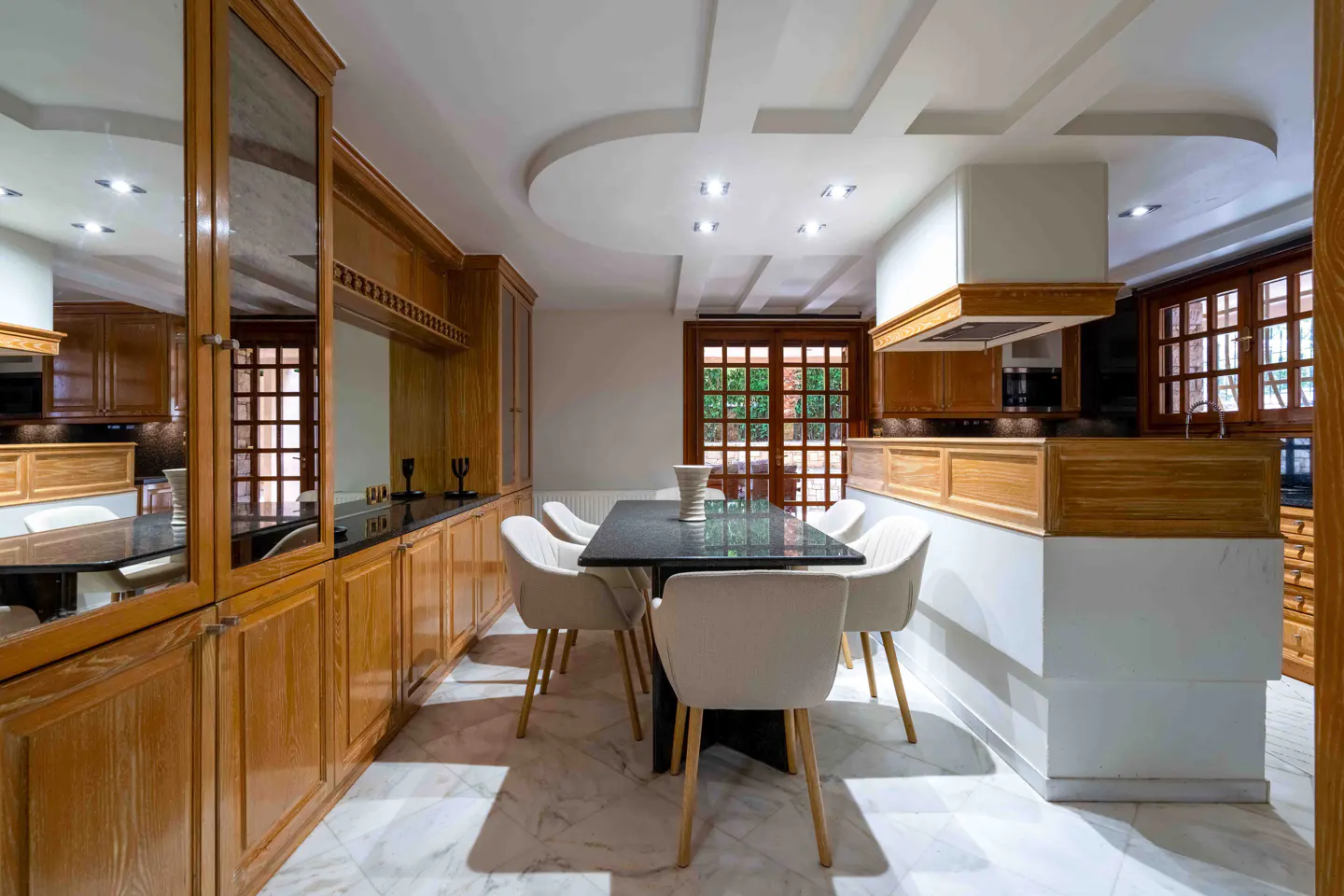 A modern kitchen and dining area with wood cabinets, a black table, and white chairs. Marble floors and a window to the outside.