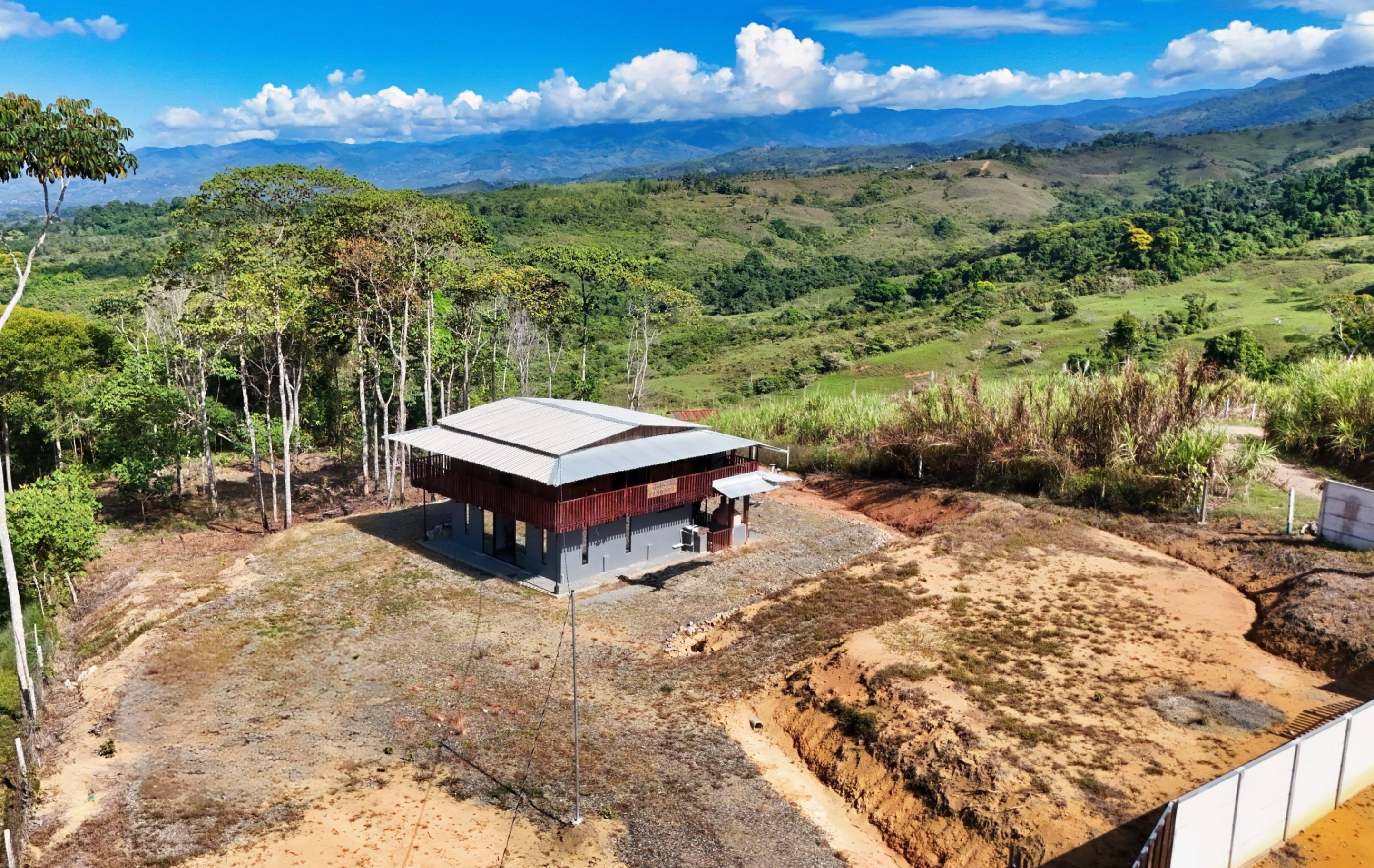 A two-story house with a gray foundation and brown siding sits on a hill surrounded by trees and mountains.