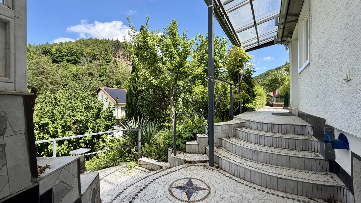 View of a home's tiled patio with steps leading to a walkway, lush greenery, and a hillside backdrop.