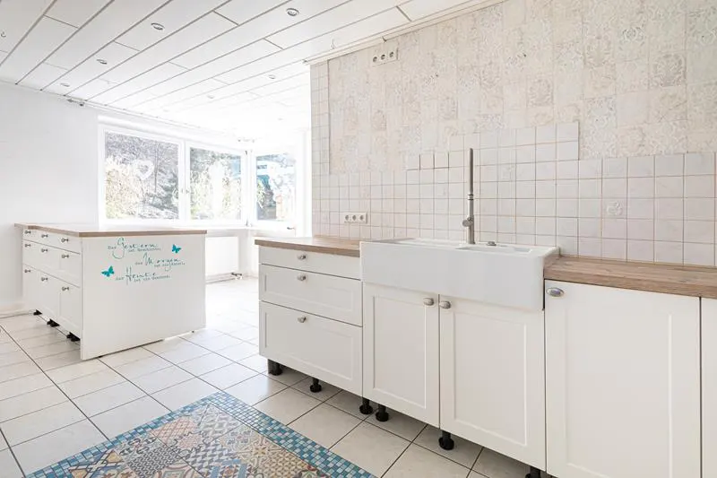 Bright kitchen with white cabinets, farmhouse sink, wood countertops, tiled walls, and a patterned rug on a tiled floor.
