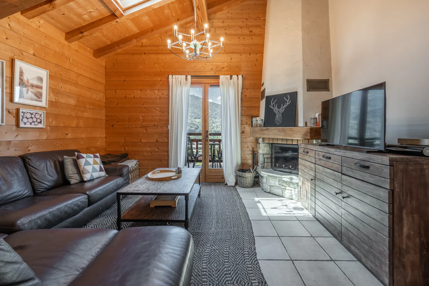 Cozy living room with wood-paneled walls, leather sofa, stone fireplace, and balcony with mountain views.