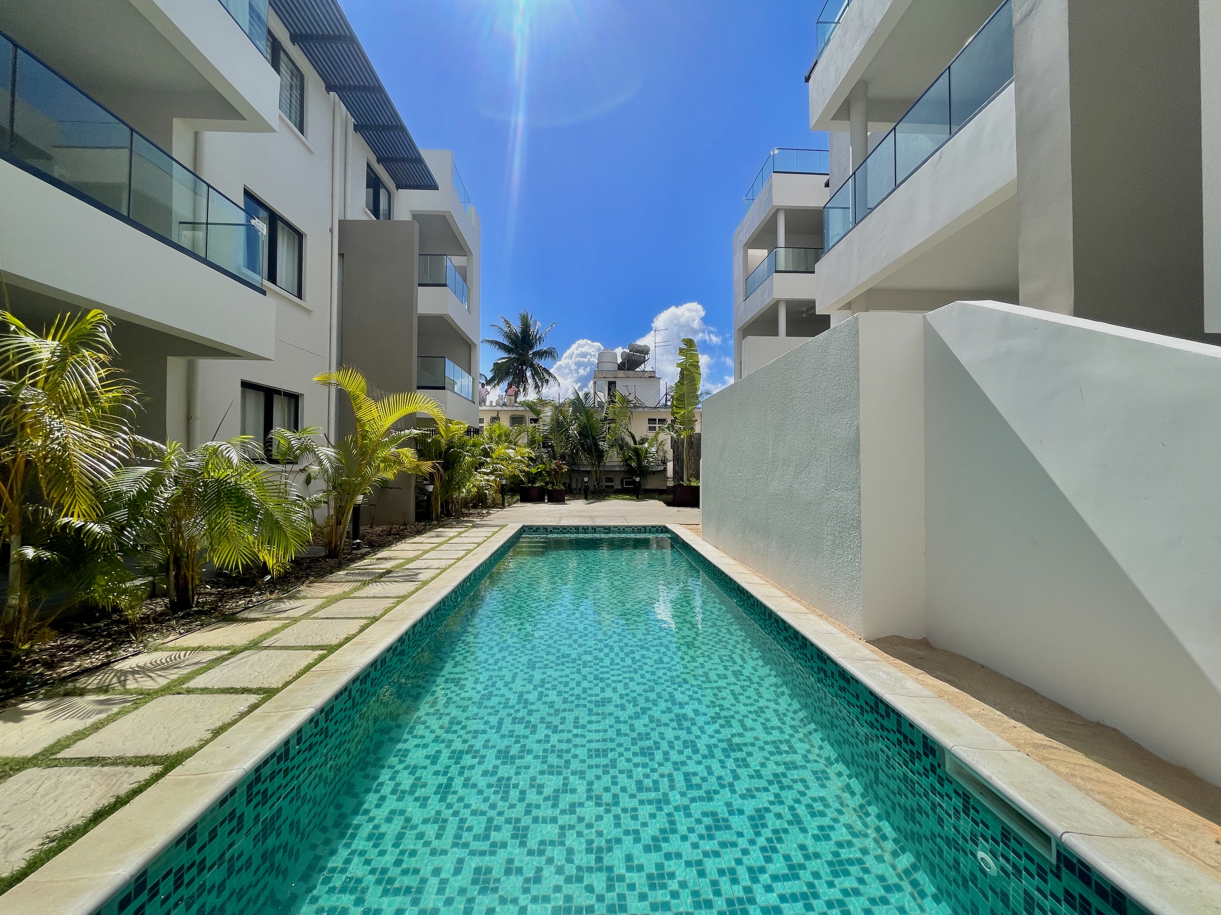 A long, rectangular swimming pool with turquoise tiles sits between two modern white apartment buildings under a blue sky. [AI-generated text]