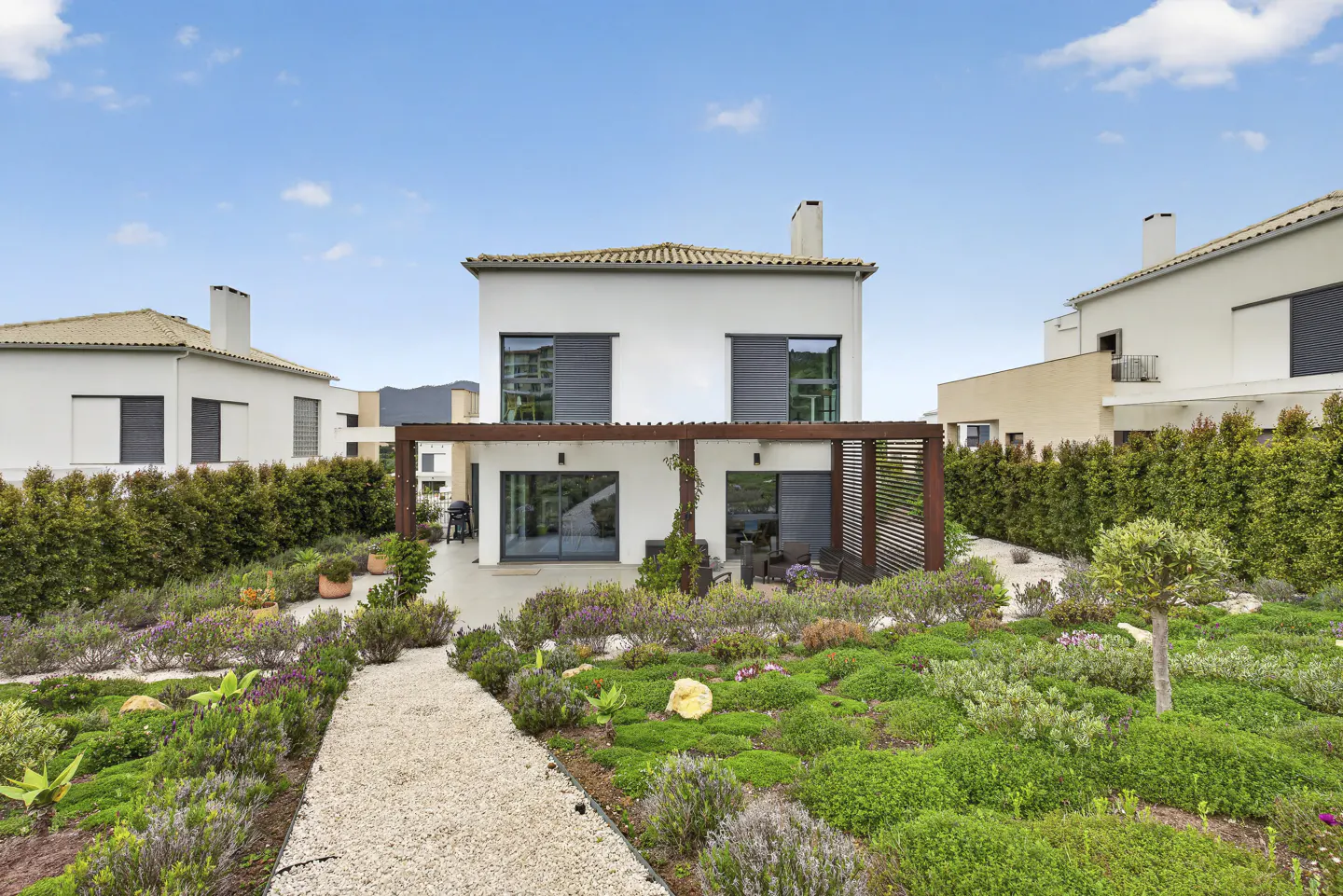 Two-story white house with a brown pergola, a stone path, and a green garden under a blue sky.