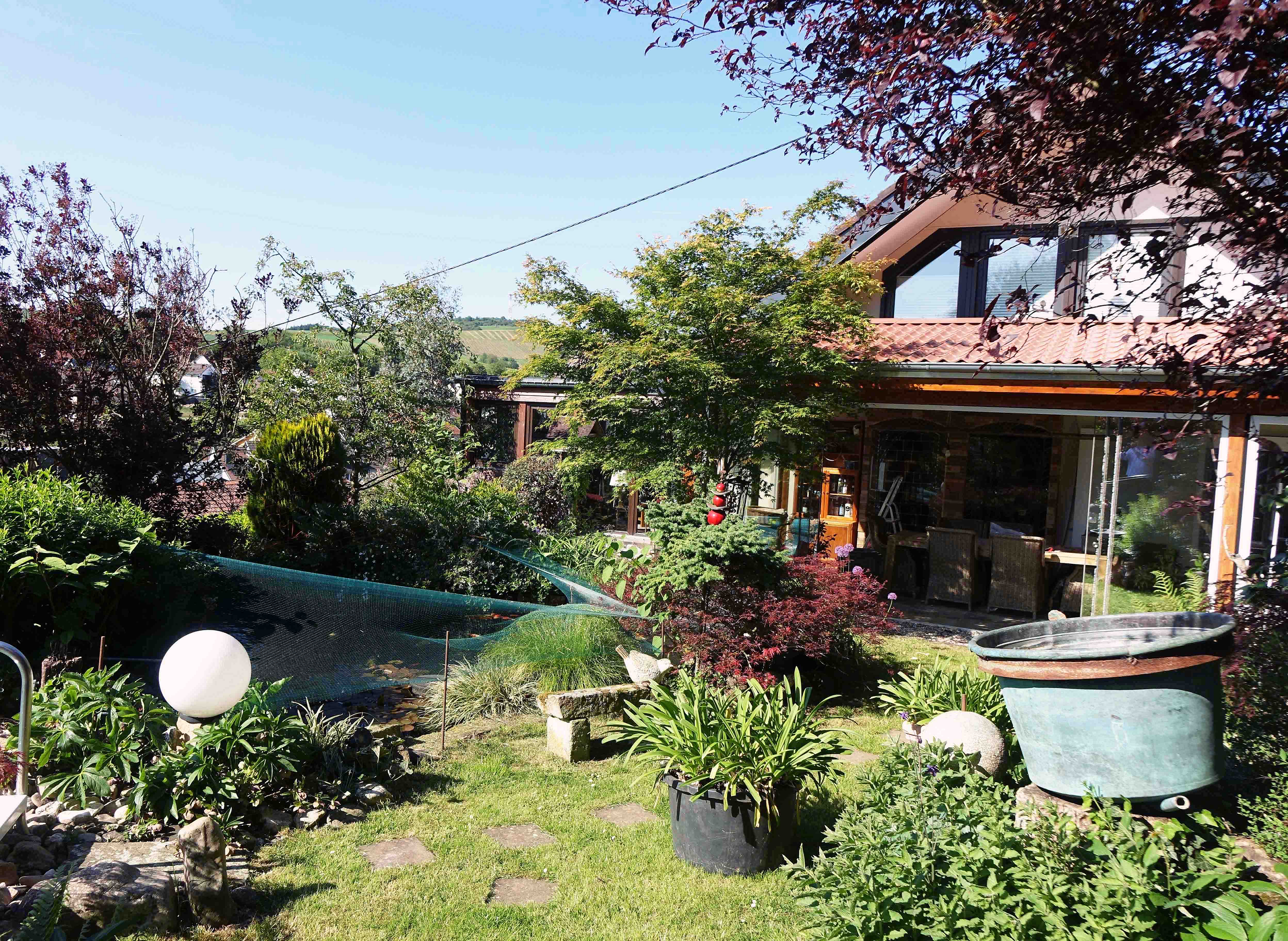 Lush garden with a pond, large copper basin, and patio area next to a house with a tiled roof under a clear blue sky.
